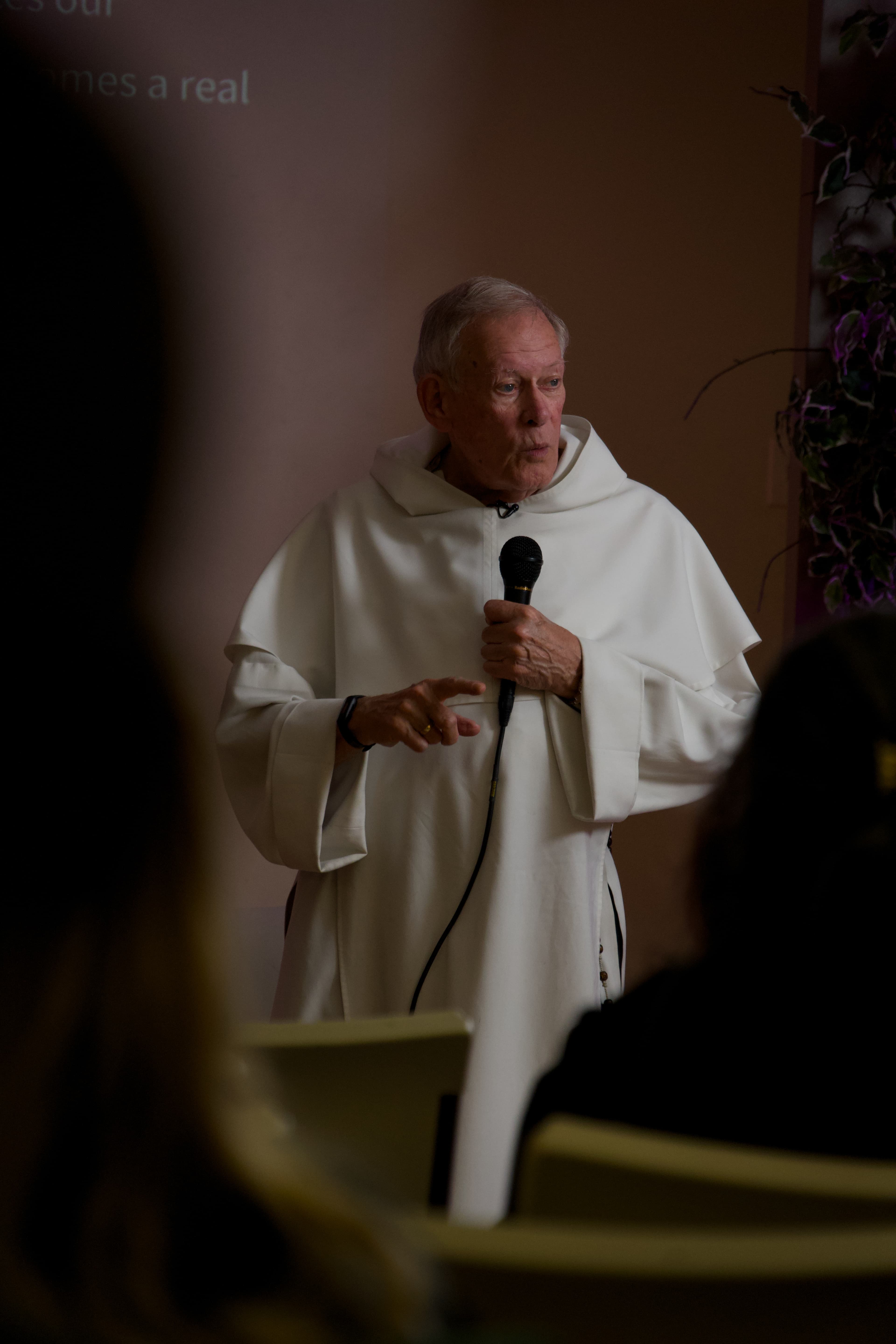 A man in a white robe speaking into a microphone, framed by the silhouettes of seated listeners