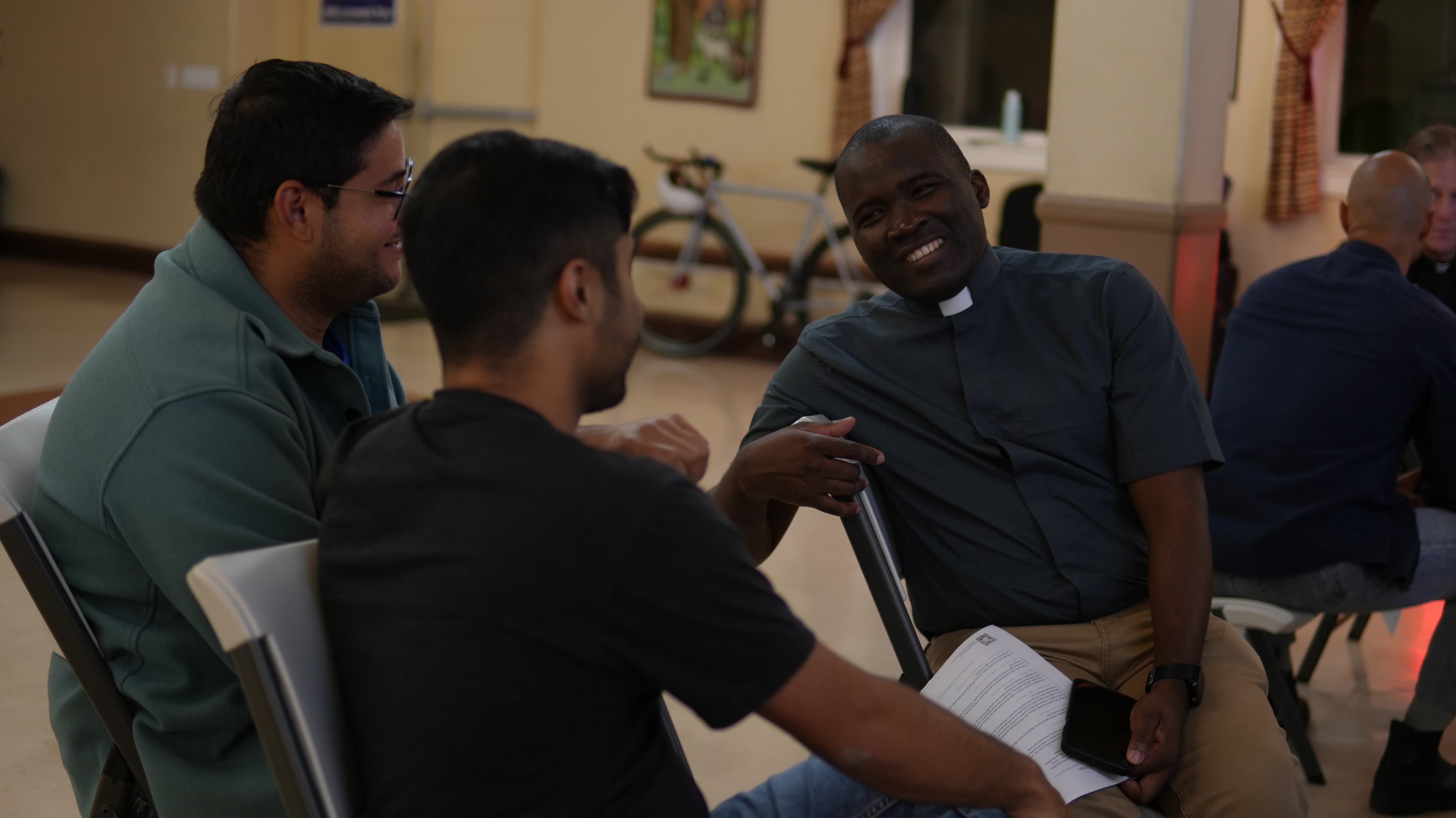 A smiling man gesturing as he talks with two other men seated nearby in a meeting room