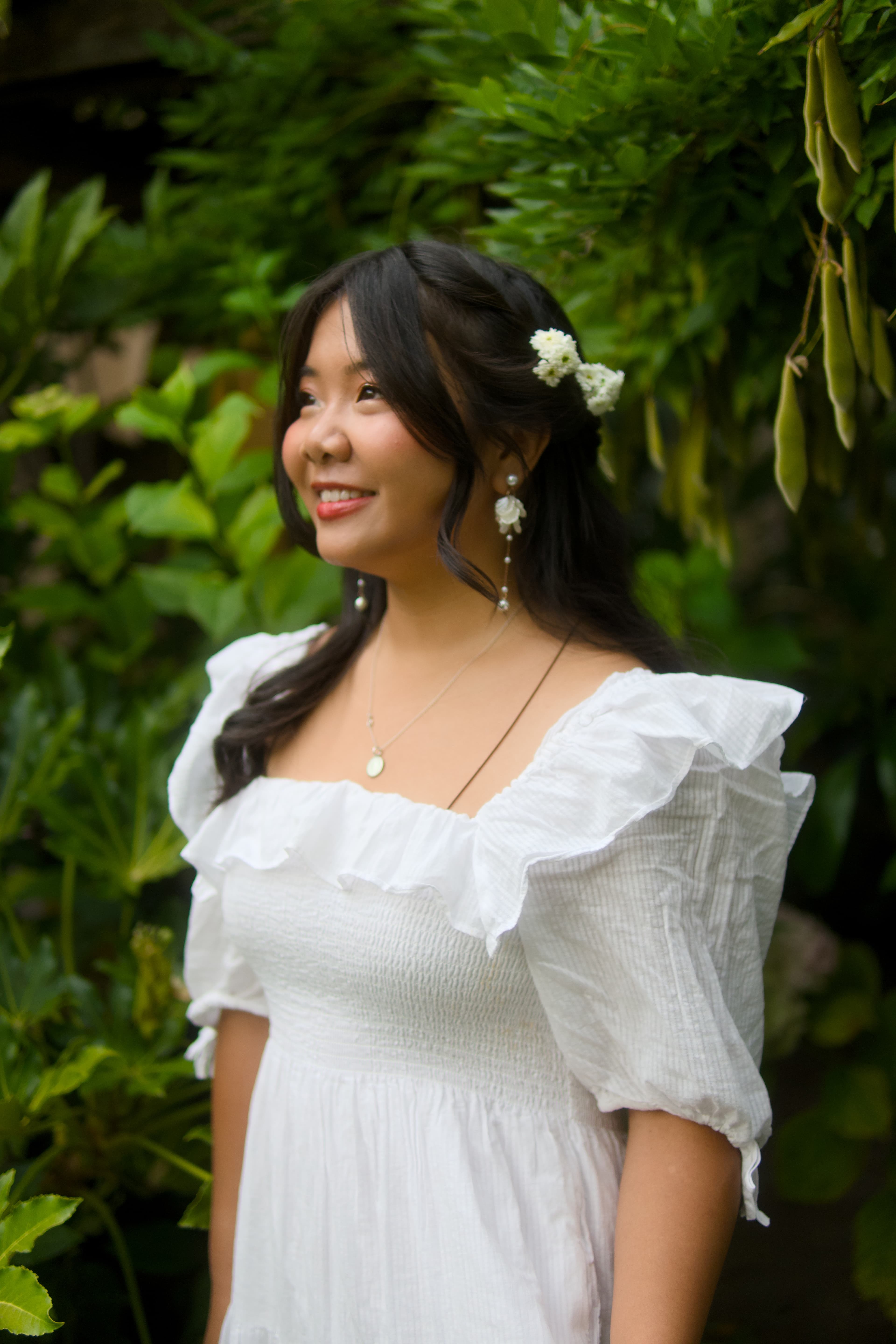 A woman in a white dress smiling beneath leafy branches, with small white flowers in her hair