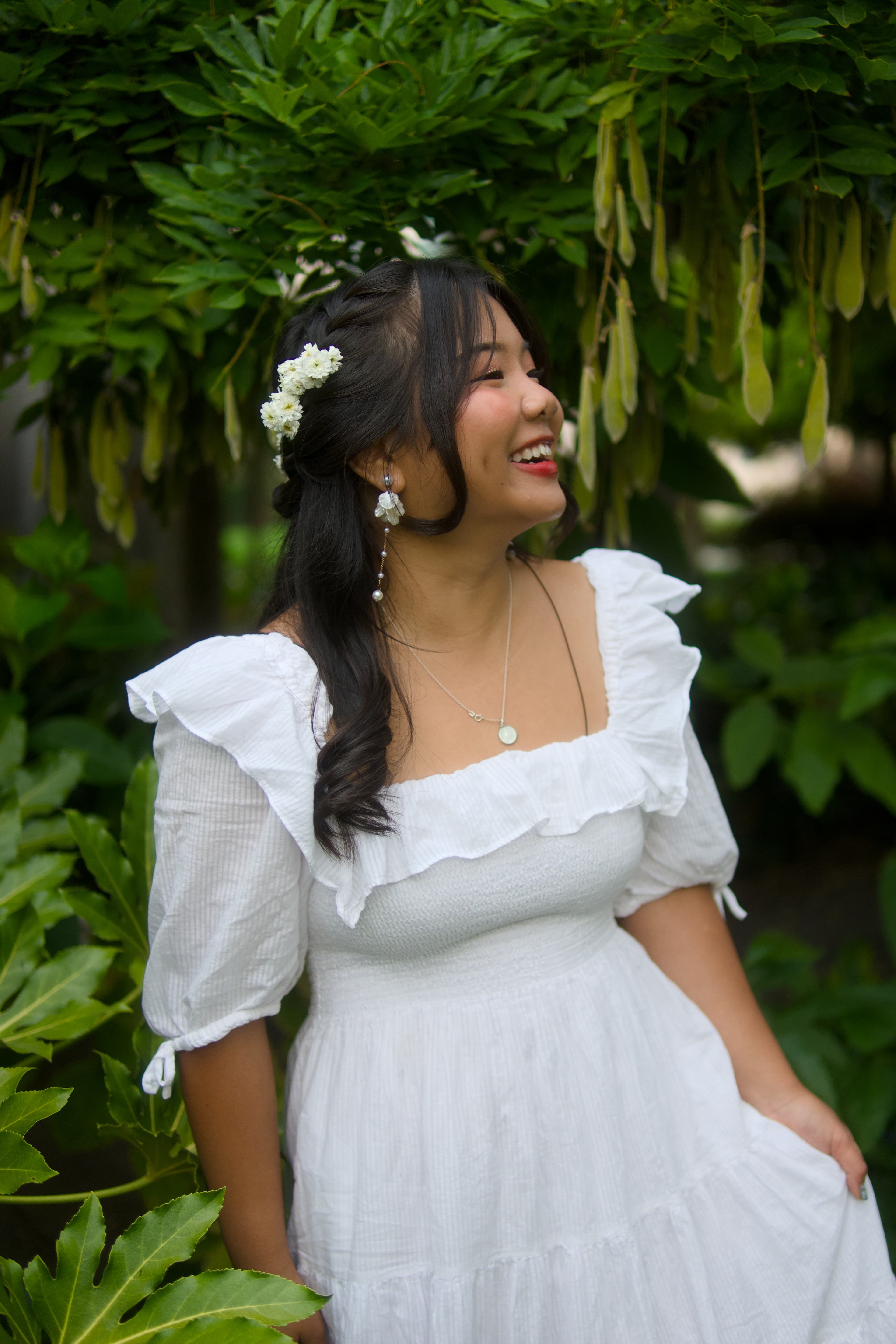 A woman in a white dress turning to the side and smiling amid lush green foliage