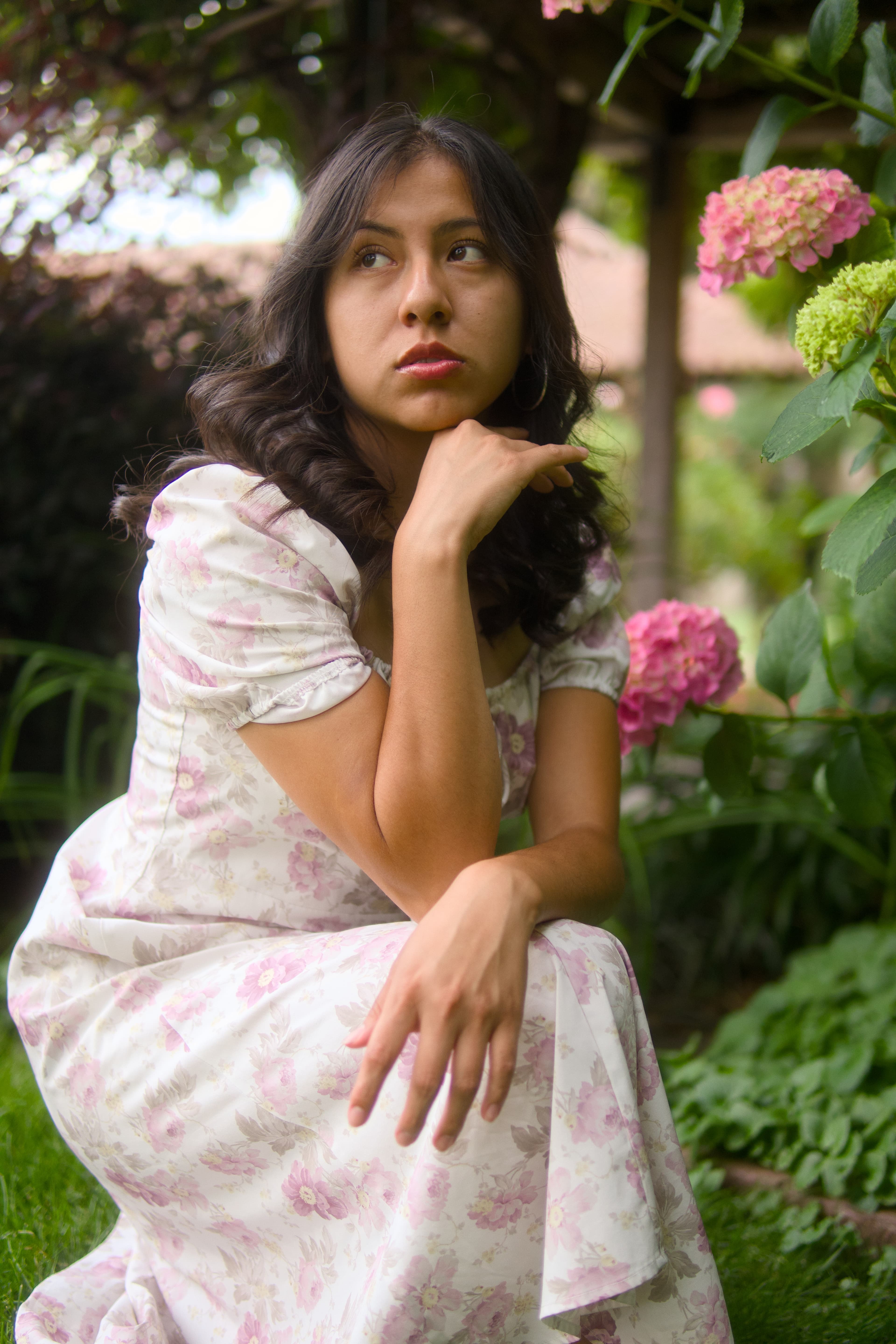 A woman in a pale floral dress resting her chin on her hand, framed by pink hydrangeas and green leaves