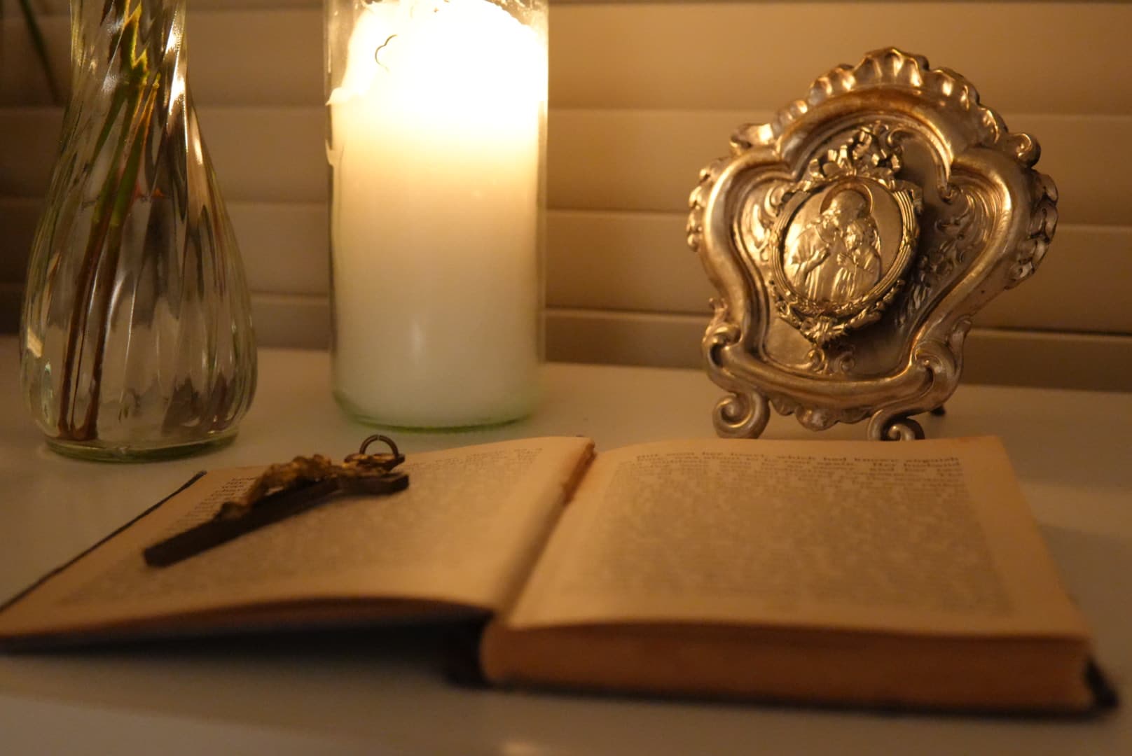 An open book and crucifix on a tabletop beside a lit candle, glass vase, and ornate frame
